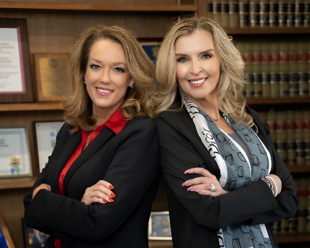 Two confident professional women smiling with arms crossed in a library setting.
